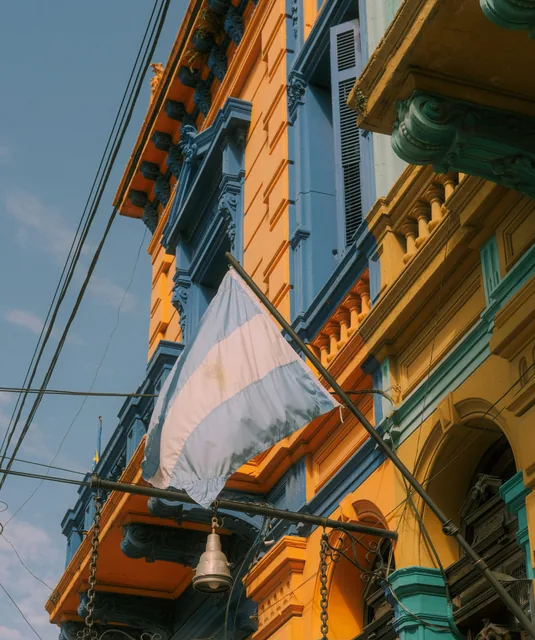 An Argentine flag hanging from a balcony on a yellow and blue building.